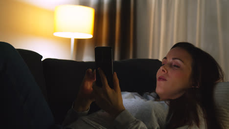 Mujer-Tumbada-En-El-Sofá-De-Casa-Por-La-Noche-Transmitiendo-O-Mirando-Contenido-En-Línea-En-El-Teléfono-Móvil-9